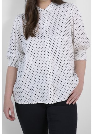 Kobieta ubrana w białą bluzkę na guziki w czarne grochy z marszczonymi rękawami 3/4, w połączeniu z czarnymi spodniami, stojąca z ręką na biodrze.