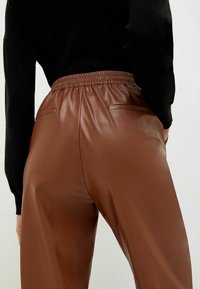 Pantaloni in faux leather marroni con cinturino elastico e due tasche posteriori, caratterizzati da una texture liscia e una vestibilità comoda.