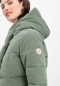Veste en duvet vert en velours côtelé avec une grande capuche, présentant une surface texturée et un accent circulaire en patch sur l'épaule.