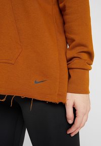 Gros plan d'une personne portant un haut marron à manches longues avec un ourlet usé et un petit logo près du bas, associé à un legging noir.