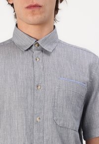 Chemise à manches courtes en tissu texturé gris clair avec un col boutonné, une poche avant avec un accent bleu et des boutons marron.
