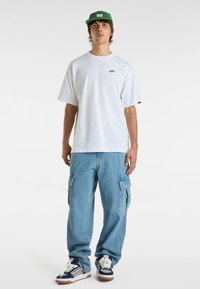 T-shirt bianca in cotone, vestibilità ampia, con piccolo logo ricamato. Pantaloni cargo azzurri chiari con tasche laterali, abbinati a sneakers blu navy e bianche. Cappellino verde.
