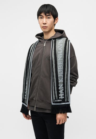 Πλεκτό κασκόλ σε γκρι, μαύρο και μπλε με κρόσσια στα άκρα. Διακοσμημένο με το γραφικό κείμενο "HAN KJØBENHAVN" κατά μήκος και μαλακή υφή.