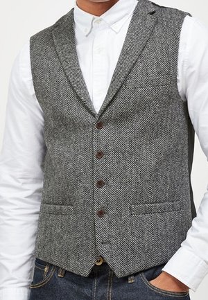 Homme portant un gilet gris à motif chevrons sur une chemise blanche à boutons, avec un jean bleu foncé, main dans la poche.