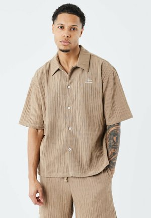 Camisa de botones de manga corta en tejido texturizado color marrón claro, con un sutil patrón de rayas y un logo blanco en el lado izquierdo del pecho.