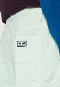 Light green denim shorts with side pockets, featuring visible stitching and a black and blue embroidered logo on the back pocket.