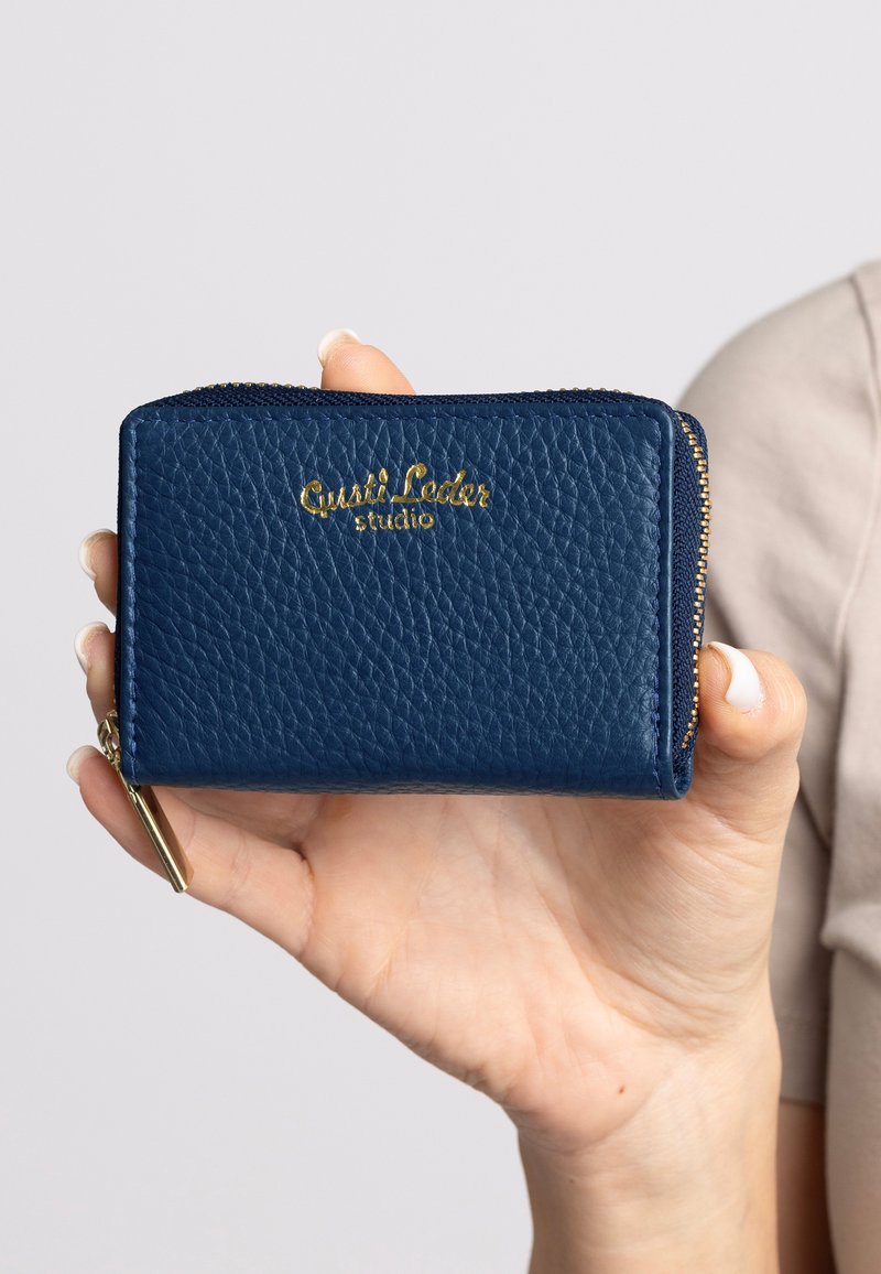 Hand hält eine kleine strukturierte dunkelblaue Leder-Brieftasche mit goldenem Reißverschluss und "Gusti Leder studio" in goldener Schrift geprägt.