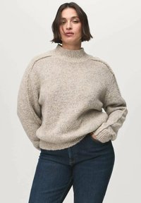 Suéter de punto gris claro con cuello alto, tejido texturizado, puños acanalados y un corte holgado, combinado con unos jeans azul oscuro.