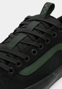 Baskets noires avec une bande latérale vert foncé, des œillets noirs et des lacets noirs passés dans une boucle centrale en tissu vert sur la languette.