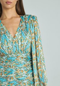 Mujer vistiendo un vestido de manga larga con un patrón geométrico abstracto en azul, verde azulado, blanco y marrón, con detalle de cintura fruncida.