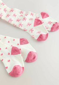 Paket mit zwei Paar Baumwollsocken. Ein Paar hat pinke Schleifen auf weißem Hintergrund, das andere hat pinke Herzen mit kontrastierenden pinken Fersen.