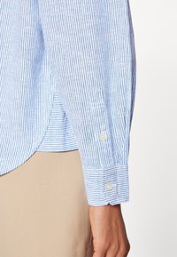Poignet de chemise à manches longues rayée bleu clair et blanc avec deux boutons blancs, portée sur un pantalon beige, sur un bras à la peau claire.