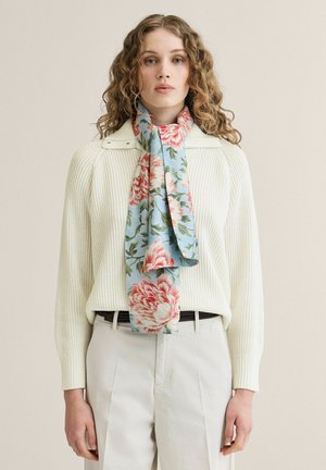 Ung kvinde med krøllet hår iført en hvid ribbet sweater, beige bukser og et blåt blomsteret tørklæde bundet om halsen.