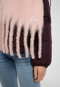 Lyserød uld tørklæde med forlængede frynser, broderet "Lee" logo. Båret over en mørk lilla dunjakke og lyseblå jeans.