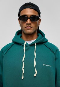 Groene hoodie met crèmekleurige trekkoorden, voorzien van een wit logo. Draagt zwarte ovale zonnebril en een zwarte baseballcap. Glad van structuur.