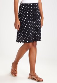 Jupe noire à pois avec un ourlet festonné, fabriquée en tissu léger. Elle présente une taille ajustée et une silhouette évasée, portée avec des sandales marron.