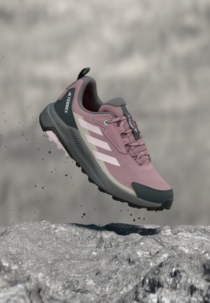 Lila-grauer Adidas Terrex Traillaufschuh schwebt in der Luft über felsigem Gelände, wobei Schmutzpartikel darunter fallen.