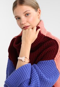 Överdimensionerad, flerfärgad stickad tröja med maroon-, rosa- och blå sektioner. Modellen bär ett ljust armband på handleden.