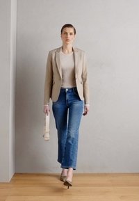 Béžový přizpůsobený blazer přes světlý žebrovaný top, v kombinaci s modrými rozšířenými džíny a béžovými lodičkami, držící světlou kabelku.