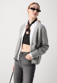 Cardigan grigio in lana su una camicia bianca con bottoni, top corto nero e jeans grigi. Gli accessori includono una sciarpa nera e occhiali da sole.