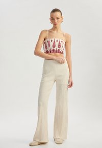 Witte crop top met rode geometrische borduursels, gecombineerd met crèmekleurige, gestructureerde wijde broekspijpen. Model staat tegen een neutrale achtergrond.