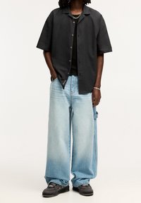 Camicia nera a maniche corte, jeans a gamba larga azzurri chiaro con sfumatura degradé, tessuto strutturato, abbinati a sneakers scure con dettagli.