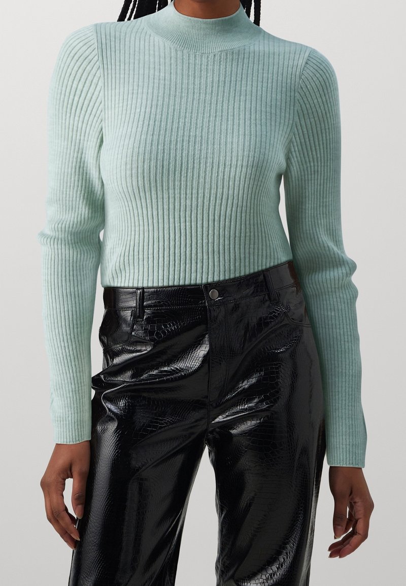 Personne portant un pull à manches longues côtelé bleu clair et un pantalon noir taille haute en simili cuir avec un motif crocodile discret.