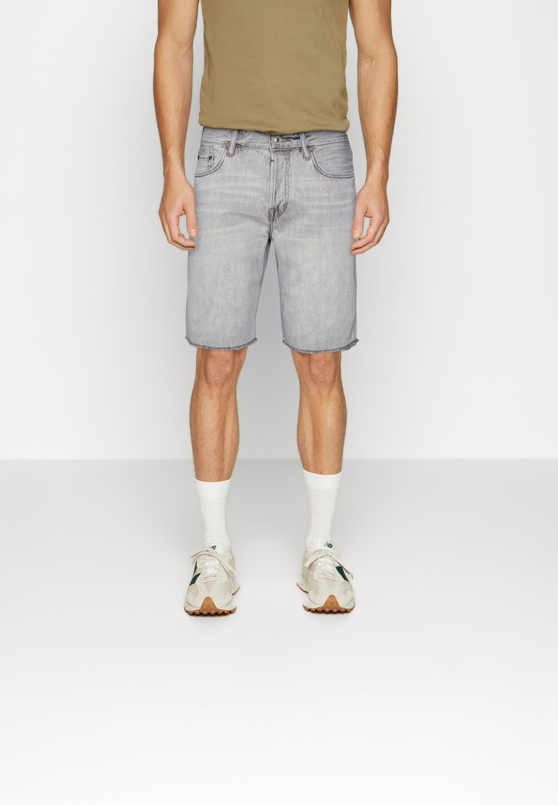 Shorts en jean gris clair avec un ourlet effiloché, associés à un T-shirt ajusté vert olive et des chaussettes de sport blanches. Les chaussures sont blanches avec des accents verts.