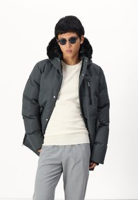 Doudoune noire avec capuche en fausse fourrure, poche zippée, et pull gris en dessous, associée à un pantalon gris clair et des lunettes de soleil.