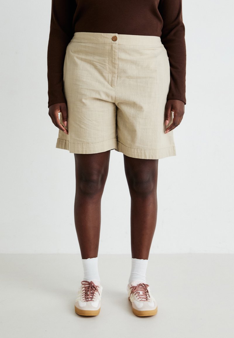 Pantalones cortos beiges con cierre de botón y textura sutil, que presentan un diseño de pierna ancha y dobladillo doblado, combinados con zapatos rosa claro y calcetines blancos.