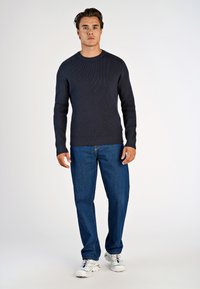 Dunkelblauer gerippter Pullover mit langen Ärmeln und Rundhalsausschnitt, kombiniert mit blauen Jeans und weißen Turnschuhen, vor einem schlichten Hintergrund stehend.