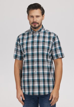 Mężczyzna w krótkim rękawie, zapinanej na guziki koszuli w kratę w odcieniach szarości, niebieskiego i czarnego, stojący na tle jednolitego jasnego tła.