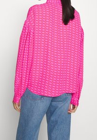 Fuksjowa koszula z długim rękawem z powtarzającym się wzorem logo, luźny krój i wysoki kołnierz. Połączona z jasnoniebieskimi dżinsami.