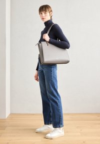 Graue strukturierte Handtasche mit schlankem Profil, goldenen Beschlägen und seitlichem Reißverschluss. Modell trägt einen figurbetonten Rollkragenpullover, blaue Jeans und weiße Turnschuhe.