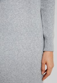 Ljust grått ulltröja med ruggade muddar, som visar en mjuk textur och slät tyg. Närbild av plagg och handdetalj.