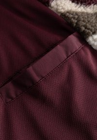 Bordó textúrált anyag, hálós mintával, egy sima bordó zsebeléssel. Simító felület enyhe fényességgel, részletek a zseb szélén.