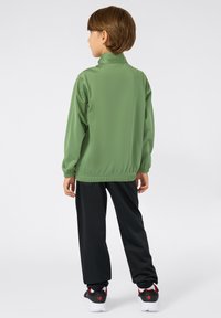 Kind met kort bruin haar staat met de rug naar ons toe, gekleed in een groene jas, zwarte broek, witte sokken en zwarte sneakers met rode accenten.