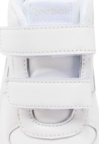 Sapatilha de couro branco com uma textura suave, apresentando tiras de Velcro ajustáveis e recortes perfurados para respirabilidade. Marca Reebok visível.