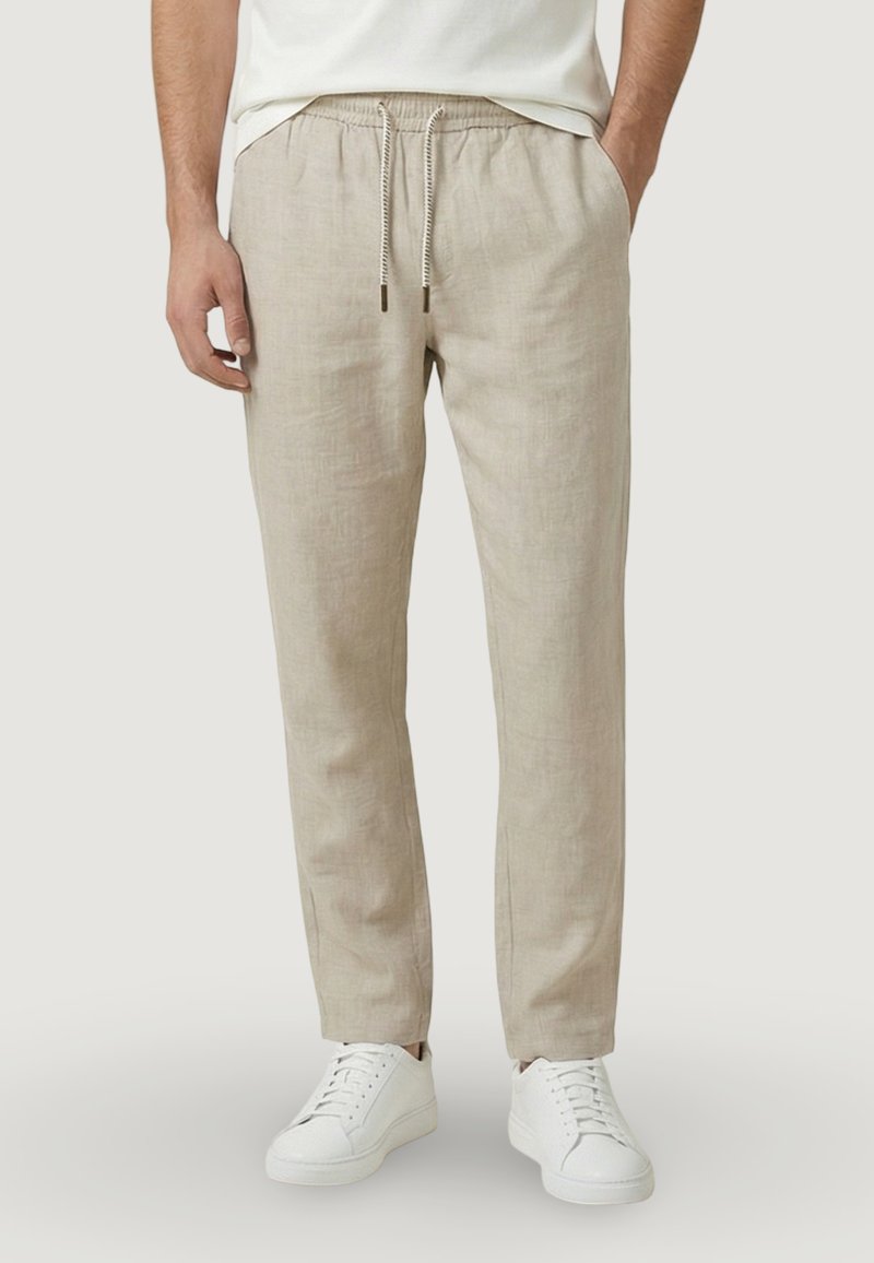 Uomo che indossa pantaloni di lino beige con coulisse e sneakers bianche basse, in piedi con le mani nelle tasche contro uno sfondo semplice.