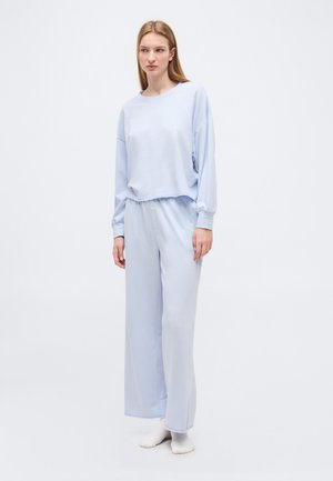 Młoda kobieta stojąca w luźnej, jasnoniebieskiej bluzce z długim rękawem i szerokich spodniach, mająca na nogach białe skarpetki na jednolitym białym tle.