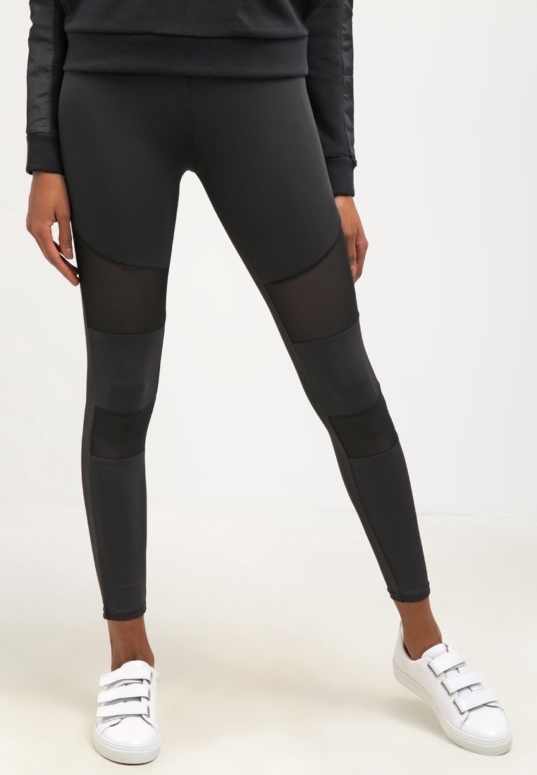 Zwarte sportlegging met meshpanelen, strak design, gladde textuur en gestikte accenten. Gecombineerd met witte sneakers met meerdere banden.