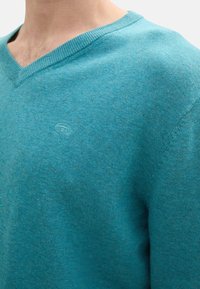 Türkiser V-Neck-Pullover aus weichem Strickstoff, mit feiner Textur und kleinem Logo auf der Brust.