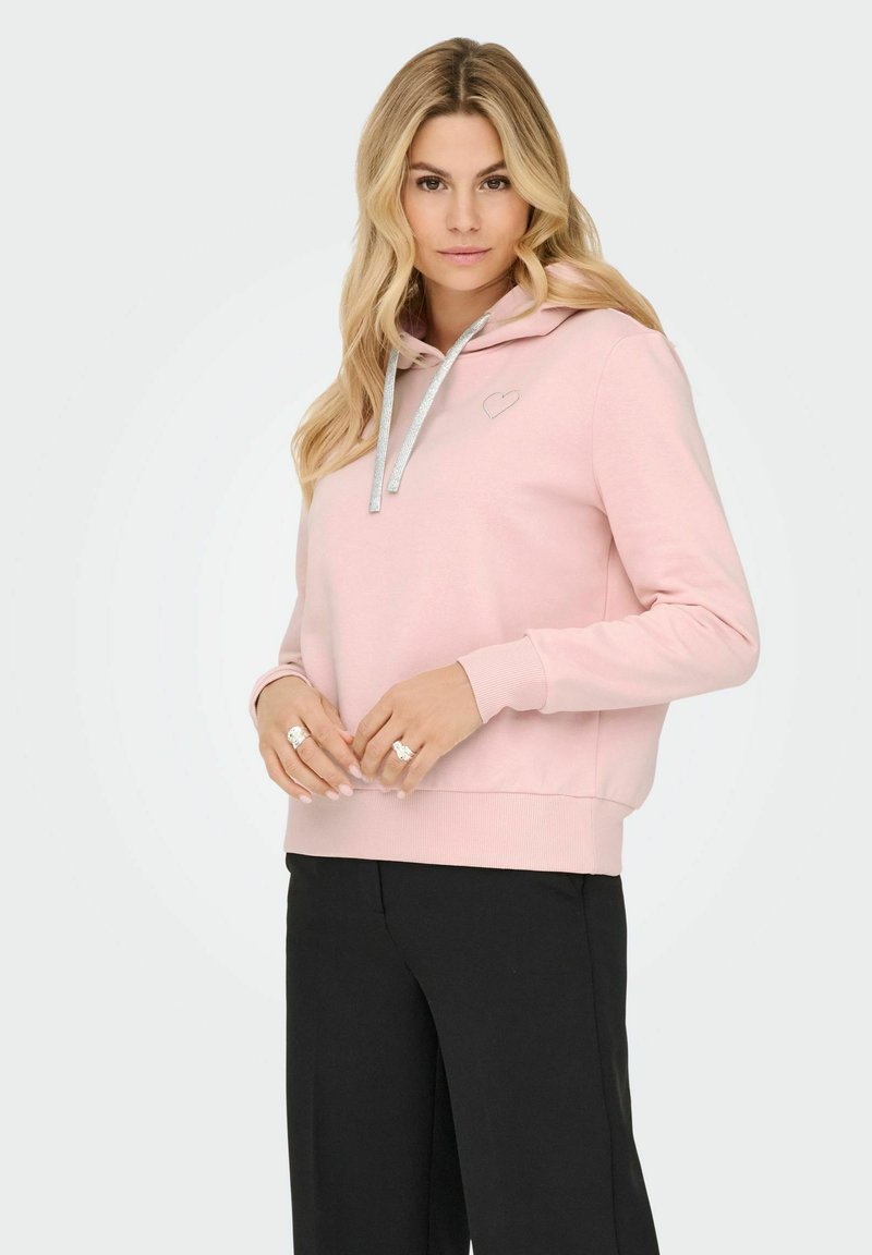 Rosa hettegenser med en sølv hjerte-logo, snorhette, lange ermer og ribbestrikket kant, kombinert med svarte bukser. Glatt tekstur.