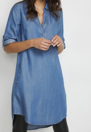 Femme portant une tunique en denim bleu avec les manches retroussées, les mains jointes devant sur un fond blanc.