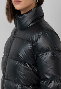 Melna pūka jaka ar augsto apkakli, izgatavota no spīdīga materiāla. Iekļauj kvadrātveida šuves un apaļo logotipa akcentu uz piedurknes.