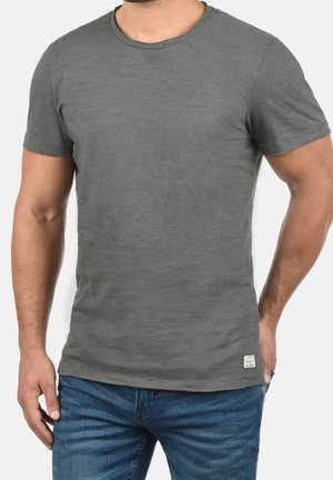 Homme portant un t-shirt gris ajusté à manches courtes et un jean bleu, debout avec une main dans la poche, sur un fond uni.