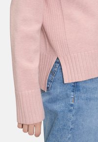 Różowy sweter dzianinowy z prążkowanymi mankietami i bocznymi rozcięciami, w zestawieniu z niebieską spódnicą dżinsową. Gładka tekstura i luźny krój są wyraźnie widoczne.