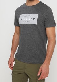 Grå bomulds T-shirt med et hvidt rektangulært grafisk print med teksten "NEW YORK HILFIGER U.S.A." Kortærmet og med rund hals.