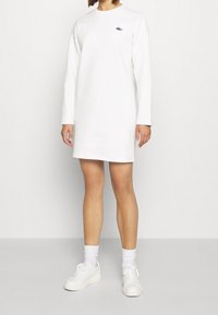 Vit långärmad klänning med rund halsringning och broderad logotyp, tillverkad av mjukt tyg, som når mitt på låret, kombinerad med vita sneakers.