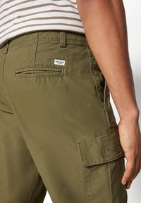 Olive green cargo pants with rear patch pocket and label; cotton material, straight cut, visible stitching details, and side pockets.